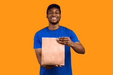 Hızlı Teslimat. Mavi şapkalı ve üniformalı Afro-Amerikan erkek kuryenin restorandan kameraya ve sarı turuncu stüdyo geçmişine el çantası taşıdığı ve verdiği ön manzara.