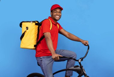 Emirleri zamanında iletiyorum. Kırmızı şapkalı ve üniformalı neşeli siyah erkek kuryenin portresi bisiklet sürüyor ve sarı termal çantalı mavi stüdyo arka planında poz veriyor.