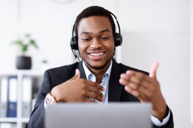 Kulaklıklı mutlu siyahi iş adamı online konferans yapıyor, ofis içi, boş yer. Çevrimiçi çalışan neşeli Afro-Amerikan yönetici, online toplantıya katılıyor, dizüstü bilgisayar kullanıyor.