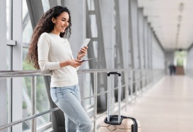 Havaalanında beklerken Smartphone kullanan güzel genç bir kadının portresi, Terminalde bavulla dururken gülümseyen Orta Doğulu bir bayanın cep telefonuna bakması, Boşluğu Kopyala