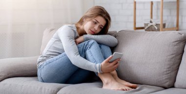 Elinde akıllı telefonuyla kanepede oturan depresif genç bir kadının portresi. Milenyum bayanı siber zorbalıktan ya da kötü bir ayrılıktan muzdarip. Ruh sağlığı konsepti