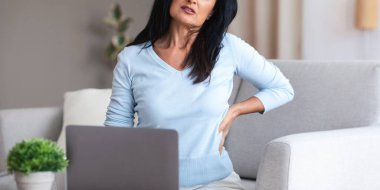 Tired Of Work. Portrait of mature lady having back pain while sitting on the couch at home, suffering from tension and acute spinal ache, feeling uncomfortable, stressed woman using laptop