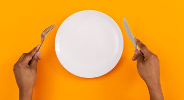 Öğle yemeği vakti. Siyah kadınlarda çatal ve bıçakla boş tabağın üst görüntüsü. Eller turuncu arka planda, panorama.