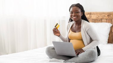 İnternetten alışveriş. Mutlu Afrikalı hamile kadın dizüstü bilgisayar ve kredi kartı ile yatakta poz veriyor, siyahi hamile bayan evde dinlenirken internetten alışveriş yapıyor, kameraya bakıyor, boşluğu kopyalıyor.