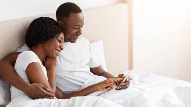 Neşeli siyah çift cep telefonu tutuyor, birlikte fotoğraf ya da video izliyorlar, evde yatakta uzanıyorlar, fotokopi çekiyorlar. Afrika kökenli Amerikalı genç adam ve kadın yatak odasında akıllı telefon kullanıyor, birlikte eğleniyorlar.