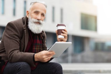Yakışıklı, gri saçlı bir adam elinde kahve bardağıyla modern dijital tablet kullanıyor, teneffüste sokakta oturuyor, fotokopi çekiyor. Yaşlı adam sabah kahvesini içip ped kullanıyor.