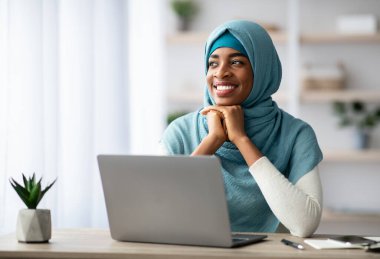 Sırasında bilgisayar başında oturan ve kafasını çevirmiş gülümseyen siyahi İslami Kadın portresi başörtülü Afrikalı Müslüman kadın İç İşleri Bakanlığı 'nda serbest çalışmanın keyfini çıkarıyor.