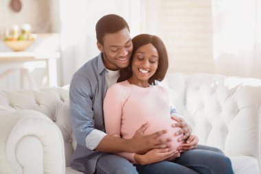 Gülümseyen anne ve baba bebek bekliyor, kanepede oturup sarılıyorlar. Kanepede kucaklaşan güzel hamile bir çift. Genç Afrikalı Amerikalı adam hamile karısını kucaklıyor.