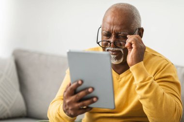 Zavallı görme yeteneği olan, gözlüklü, dijital tablet kullanan ve evde koltukta oturup haberleri okuyan yaşlı siyahi bir adam. Göz Sağlığı Sorunları Konsepti. Seçici Odaklanma