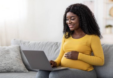 Smiling black pregnant lady using laptop at home, sitting on couch at living room, empty space. Happy african american expecting woman surfing on Internet, reading blog about maternity or shopping