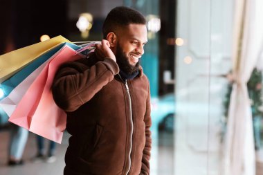 Tüketim kavramı. Afro-Amerikan bir adamın alışveriş merkezinin vitrinine bakmasından memnun oldum. Elinde alışveriş poşetleri tutan gülümseyen siyah adam, fotokopi alanı, bulanık arka plan. Perakende ve Satın Al