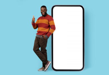 Çevrimiçi tanıtım. Neşeli siyahi adam Big Blank Smartphone 'un yanında duruyor ve başparmağını kaldırıyor, neşeli Afrikalı Amerikalı erkek cep telefonuna yaslanıyor Mockup İçin Beyaz Ekran, Tam Uzunluk