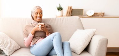 Evdeki kanepede kahve içen tesettürlü mutlu genç bir kadının tam boy portresi. Geleneksel türbanlı Afrikalı kadın sıcak bir içecekle rahat koltukta dinleniyor, hafta sonu sabahı dinleniyor.