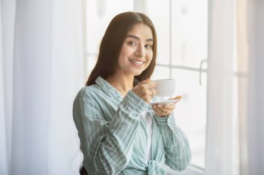 Lovely young Indian woman having coffee break in front of window at home. Charming millennial lady relaxing, enjoying lazy weekend morning with hot drink, smiling at camera