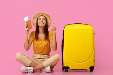 Pasaportu ve uçuş biletleri olan mutlu genç bir kadın sarı bavulun yanında oturuyor, gözlerini kapatıyor ve parmaklarını çaprazlıyor, tatil için bekliyor, pembe stüdyo geçmişi, kopyalama alanı.