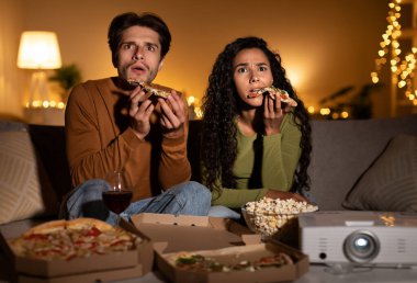 Korku filmi seyreden ve evde sinema projektörü kullanarak pizza yiyen çift geceleri oturma odasındaki koltukta oturuyor. Aile Eğlencesi ve Hafta sonu Eğlencesi Konsepti