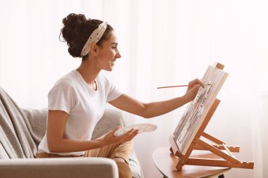 Koltukta oturup resim yapan, palet, fırça ve sehpa kullanan mutlu genç bir kadın. Yaratıcı kadın yeni hobisini seviyor, hayatı resmediyor, mekanı kopyalıyor. Eğlence konsepti