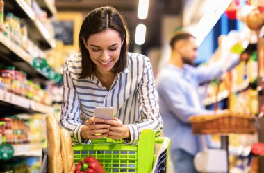 Gülümseyen Bayan Müşteri Market Alışverişi yaparken Süpermarkette Akıllı Telefon Yürüyüşü yapıyor. Seçici Odaklanma. Markette Bakkal Alışveriş Uygulaması Kullanan Kadın Telefonla Yemek Pişiriyor