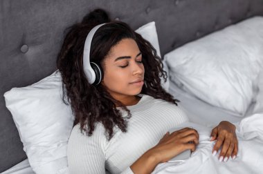 Dinlen ve rahatla kavramı. Yatakta uzanmış, müzik, ses kitabı, podcast, uyku meditasyonu ve kablosuz kulaklıkla huzurlu zihnin keyfini çıkaran sakin zenci kadın portresi. Zen benzeri