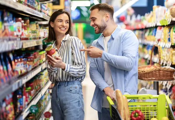Market alışverişi. Genç, gülümseyen bir çift marketten alışveriş yapıyor. İki Alıcı Alışveriş Arabasıyla Koridorda Duruyor Yiyecek Ürünlerini Seçiyor ve Gülüyor