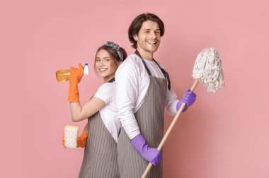 Profesyonel Temizlik Servisi. Gülümseyen adam ve kadının portresi. Ev aletleri ellerinde. Pembe stüdyonun arka planında sırt sırta duruyorlar.