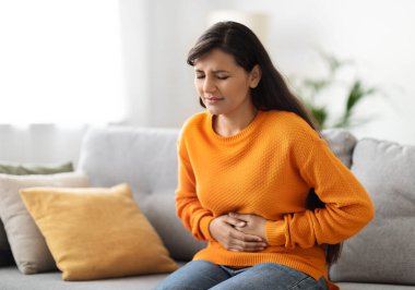 Endişeli genç Hintli kadın mide ağrısı çekiyor. Evdeki kanepede oturuyor. Kadın karnını tutuyor, karın ağrısı ve rahatsızlığı var, regl oluyor, boşluğu kopyalıyor.