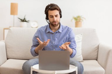 Kulaklıklı adam dizüstü bilgisayarla konuşuyor video görüşmesi yapıyor evdeki koltukta uzaktan çalışıyor. Uzak İletişim, Çevrimiçi İş Kursu Eğitimi Konsepti