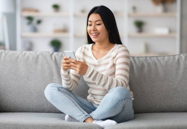 Genç Asyalı kadın evde koltukta dinlenirken akıllı telefon kullanıyor, güzel Koreli kadın koltukta cep telefonuyla oturuyor, arkadaşlarıyla mesajlaşıyor ya da oturma odasında dinlenirken yeni uygulamalara göz atıyor.