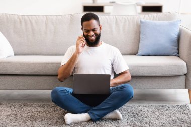 Telefonda konuşan siyahi serbest çalışan adam dizüstü bilgisayarını kullanıyor ve evde yerde oturuyor. Uzak Çalışma ve Uzak Mobil İletişim, Serbest Kariyer ve Modern Yaşam Tarzı