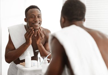 Memleketteki banyoda aynanın yanında tıraş olduktan sonra pürüzsüz bir yüz derisine dokunan mutlu siyah adam. Erkek yüz bakımı, Sabah Güzelliği Rutini ve Kişisel Bakım Kozmetik Konsepti