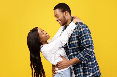 Boş zaman, hafta sonu, randevu ve romantizm. Gülümseyen Afrikalı genç Amerikalı erkek ve bayan eğleniyor, sarılıyor ve birbirlerine bakıyorlar, sarı arka planda izole edilmiş, stüdyo çekimi, profil, kopyalama alanı, kesme