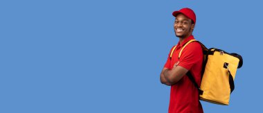 Teslimat Servisi. Kırmızı üniformalı ve termo çantalı özgüvenli siyah erkek kurye portresi katlanmış kollarla poz veriyor ve mavi stüdyo arka planında izole edilmiş kameraya bakıyor.