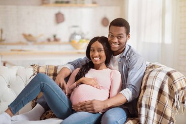 Evdeki kanepede kucaklaşan güzel hamile çift. Genç Afro-Amerikalı adam, mutlu hamile karısını evde kucaklıyor. Gülümseyen anne ve baba bebeklerini bekliyor.