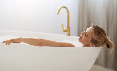 Rahatlamış sarışın kadın evde sıcak köpüklü aromatik banyodan hoşlanıyor, küvette kapalı gözlerle, yan gözle yatıyor, yakın çekim fotoğrafı, fotokopi odası. Ev spası, vücut bakımı konsepti.
