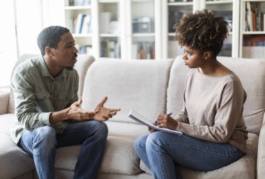 Young african american woman therapist taking notes on her upset patient at therapy session, frustrated black man having conversation with psychologist, sharing feelings, counselor office interior