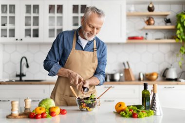 Mutlu kıdemli adam mutfakta sağlıklı sebze salatası hazırlıyor baharatlı yemek, önlüklü yaşlı beyefendi gülümsüyor bowlinge tuz ekliyor, evde vejetaryen yemekleri pişiriyor, bedava yer açıyor.