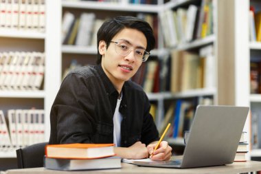Araştırma Projesi üzerinde çalışıyorum. Kütüphanede dizüstü bilgisayar kullanan, not defterine yazan ve kameraya bakan Asyalı bir erkek öğrencinin portresi.