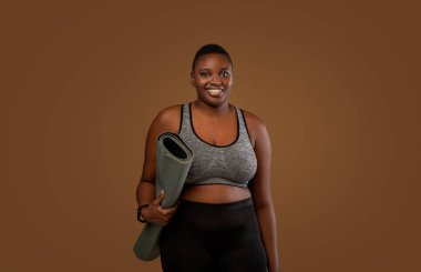 Antrenman için hazır. Sütyenli ve taytlı Afrikalı Amerikalı bir kadının portresi spor salonuna gidiyor, elinde rulo minderle kahverengi stüdyo arka planında dikilip kameraya poz veriyor.