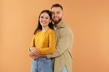 Stüdyoda turuncu arka planda kameraya gülümseyen kız arkadaşına sarılan mutlu beyaz bir adam. Millennial aile çifti fotoğraf çektirmek için kucaklaşıyor. Aşk ve romantik randevu, Sevgililer Günü