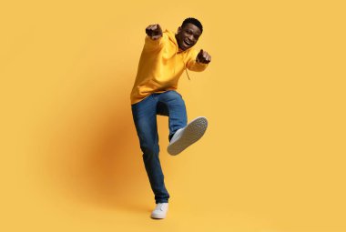 Sarı stüdyo arka planında parmak uçlarını ve ayakkabı ayakkabılarını gösteren şık giysili mutlu, kıvırcık, genç, siyah bir adam, sanki bir şey ya da biri, kopyalama alanı gibi. İnsan jestleri kavramı