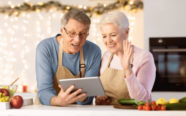 Mutlu yaşlı çift Noel arifesinde mutfakta dikilirken tablet kullanıyor. Uzaktan Xmas Kutlama Online. Daha Yaşlı Eşler Uzak Aile Kullanımı Bilgisayarıyla Sohbet Ediyor