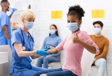 Başarılı Coronavirus aşısı. Covid-19 Aşı Aşısı 'ndan sonra hastanede başparmak hareketi yapan mutlu siyah kız. Corona Virüs Koruma ve Bağışıklık Sistemi. Seçici Odaklanma