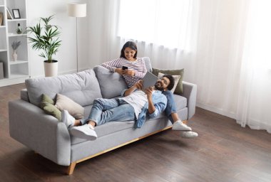 Yüksek açı, rahat, neşeli, modern, modern, doğulu aşıkların rahat oturma odasındaki kanepede dinlenen erkek ve kadınların görüntüsü. Evdeki aletleri kullanıyorlar, sosyal medyada kayıyorlar, çevrimiçi okuyorlar, uzayı kopyalıyorlar.