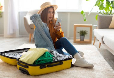 Şok edici seyahat haberleri okuyan genç bir kadın ya da evinde bavulunu hazırlarken akıllı telefondan turizmle ilgili olumsuz bir mesaj. Uçuşun iptali, covid salgını sırasında seyahat sorunları