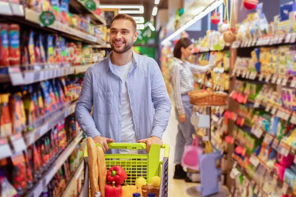 Market alışverişi. Gülümseyen Sakallı Adam süpermarkette rafların arasında alışveriş arabasıyla yürüyor. Mağazadan sağlıklı yiyecek alan mutlu adam, arka plandaki kız arkadaşı...