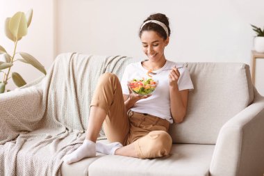 Genç ve güzel, siyah saçlı bir bayan evdeki kanepede oturuyor, renkli taze sebze salatası yiyor ve gülümsüyor, sağlıklı bir kadın vitamin dolu yiyeceklerin tadını çıkarıyor, fotokopi odası. Yemek, diyet, sağlıklı yaşam tarzı.