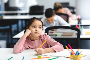 Eğitim ve Öğrenme Konsepti. Yorgun ve sıkılmış küçük Asyalı kızın portresi. Okulun masasında oturuyor, not defterine yazıyor ve düşünüyor, pencereye bakıyor, el üstünde dinleniyor.