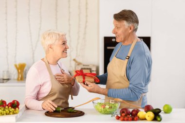 Aged Husband Giving Birthday Gift To Wife Celebrating Family Holiday Or Anniversary In Modern Kitchen At Home. Holidays Celebration, Gifts And Presents, Celebratory Dinner Concept.