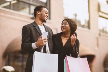 İndirim indirimi, internetten alışveriş ve online uygulamadan Kara Cuma. Gülümseyen Afro-Amerikalı erkek ve kadın renkli çantalı ve telefonlu. Yürüyüş, açık hava, fotokopi alanı.