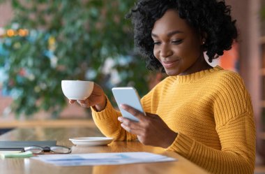Young afro girl having morning coffee at cafe alone, using phone, close up, empty space